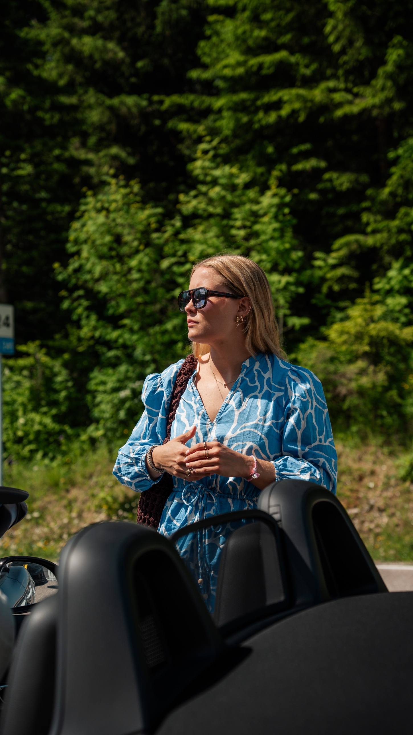 Beifahrerin mit Sonnenbrille neben einem offenen Roadster vor Wald