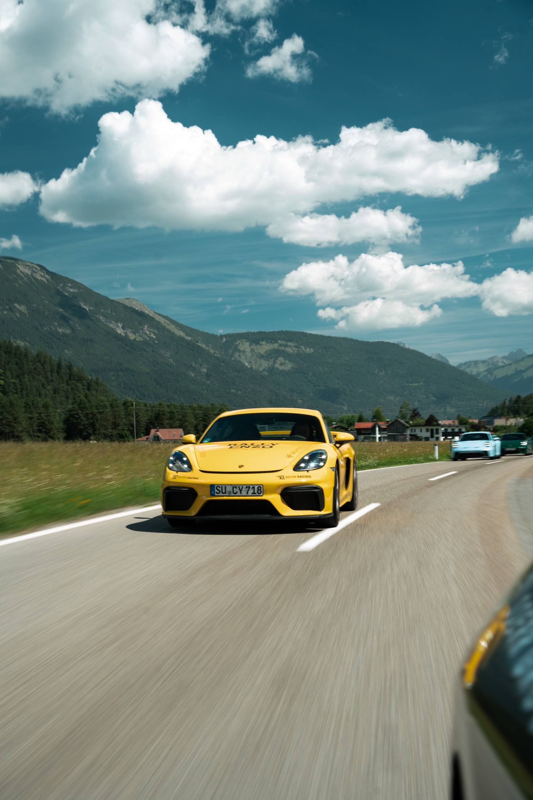 Gelber Porsche Cayman mit RALLY-ENZO-Beklebung vor Alpenpanorama, dahinter weitere Teilnehmer
