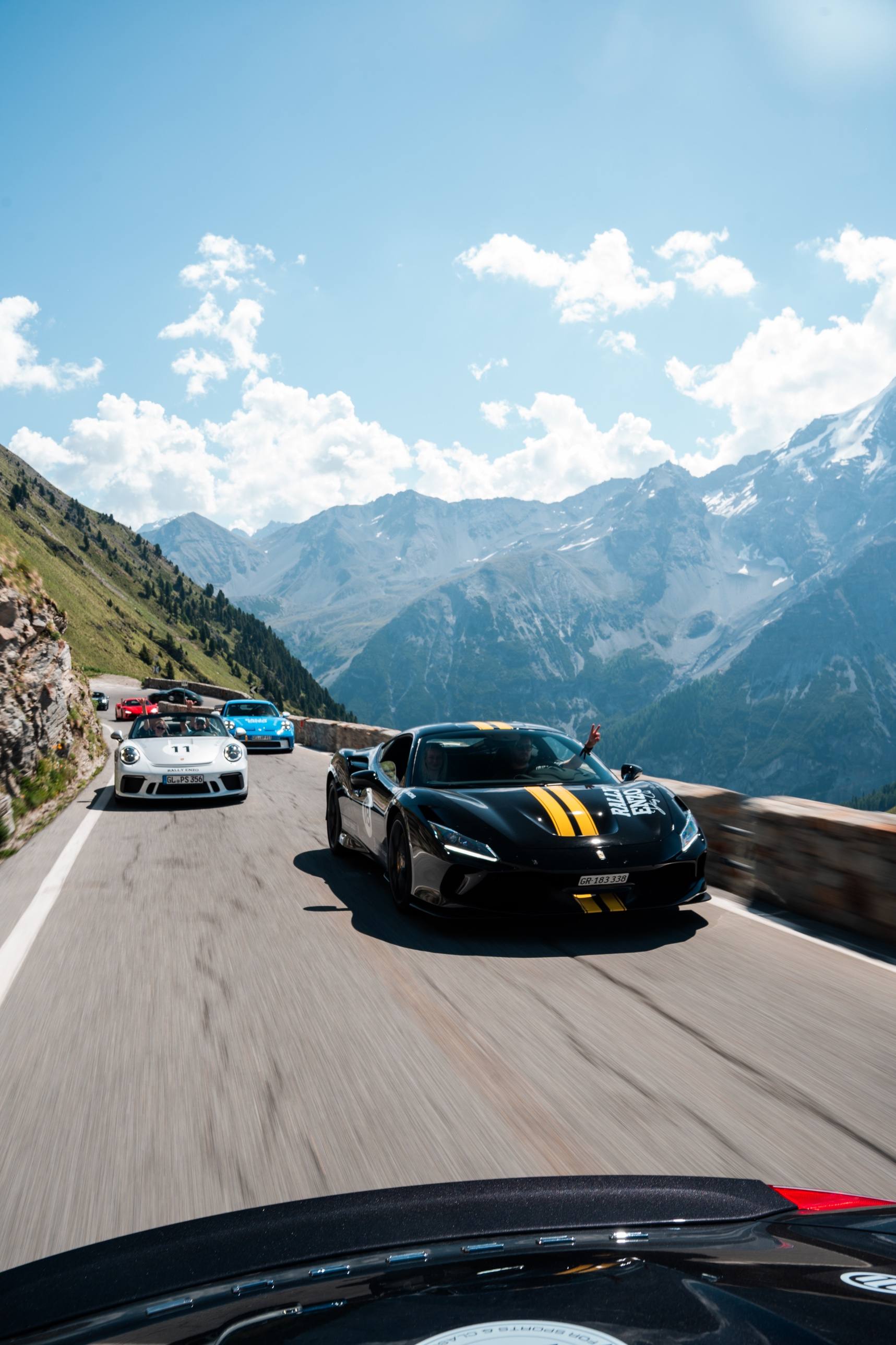 Kolonne aus schwarzem Ferrari mit gelben Streifen, Porsche Speedster mit Startnummer 11 und blauem GT3 auf einer Passstraße vor Gletscherbergen